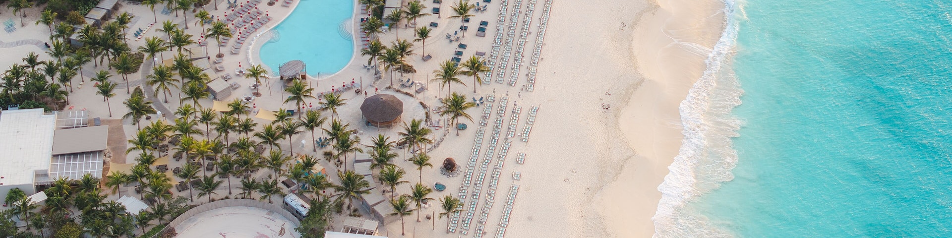 Aerial view of turquoise waters meet the sandy shore beside a resort with winding pools and palm trees, Alice Town, Bimini, The Bahamas.