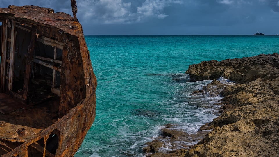 A weathered shipwreck on a rocky coastline with turquoise waters and a distant ship.