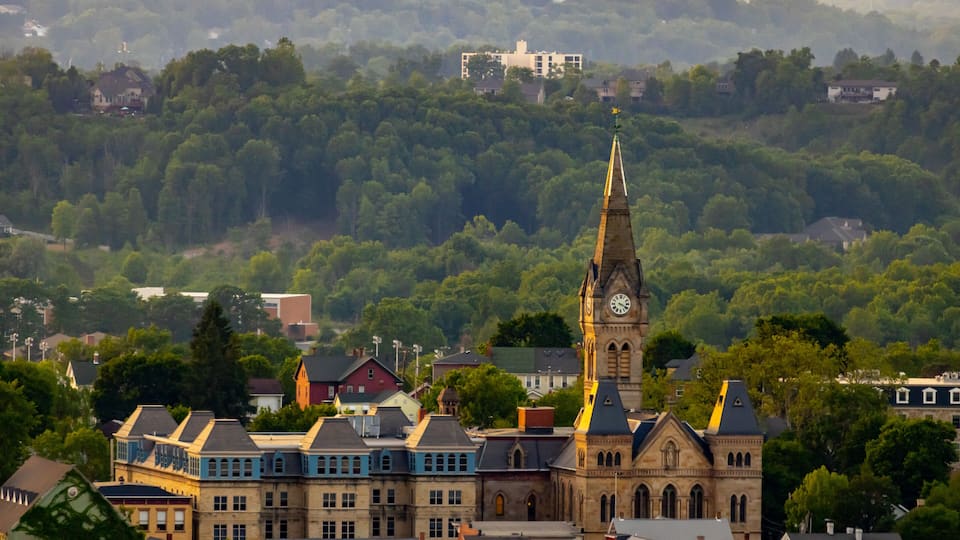 A town with a prominent church featuring a tall steeple and clock tower, surrounded by colorful buildings and dense forested hills in the background.