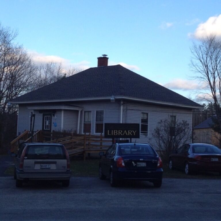 A library building with a sign, parked cars, and a wooden fence.