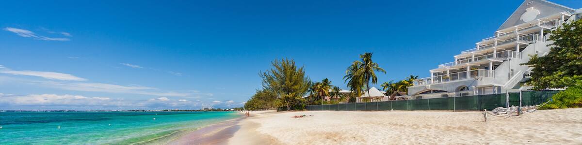 A beach with clear turquoise water, a sandy shore, and a modern building with balconies.