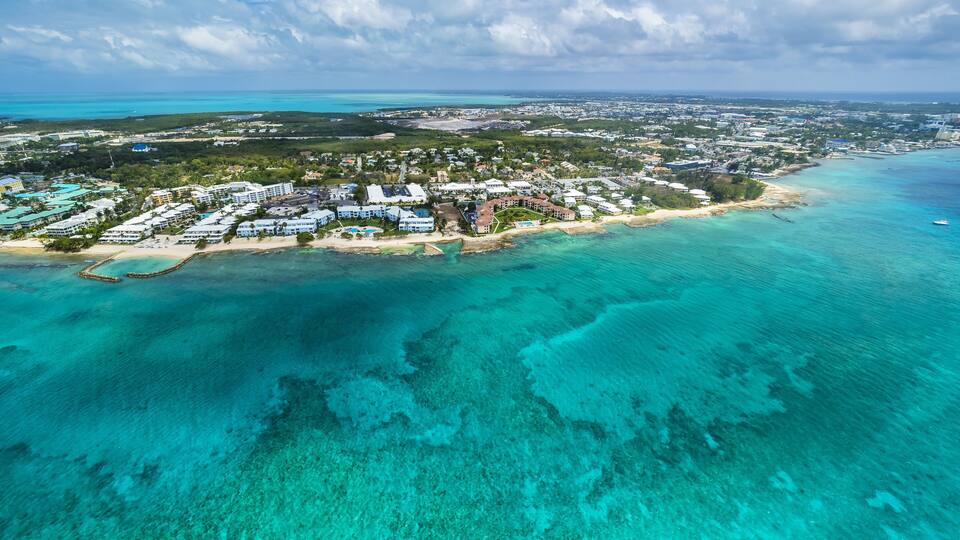 An aerial view of a coastal resort with clear turquoise waters, a sandy beach, and a mix of residential and hotel buildings.