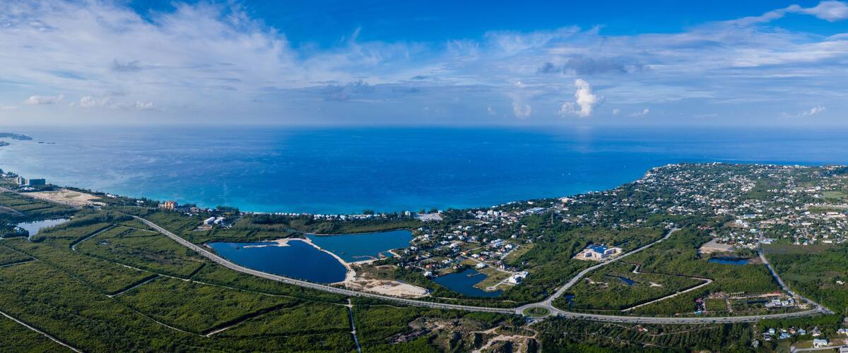 An aerial view of a coastal town with a large body of water, green hills, and a highway.