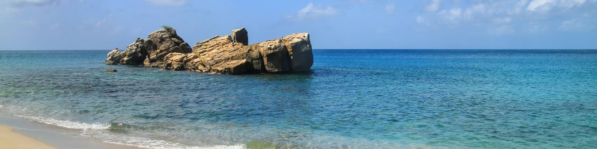 A sandy beach with clear turquoise water and a rocky outcrop in the distance.