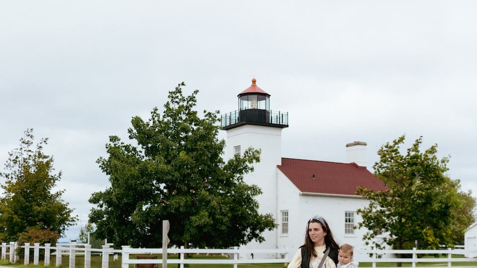 A woman and child walking on a gravel path with a lighthouse and white fence in the background.