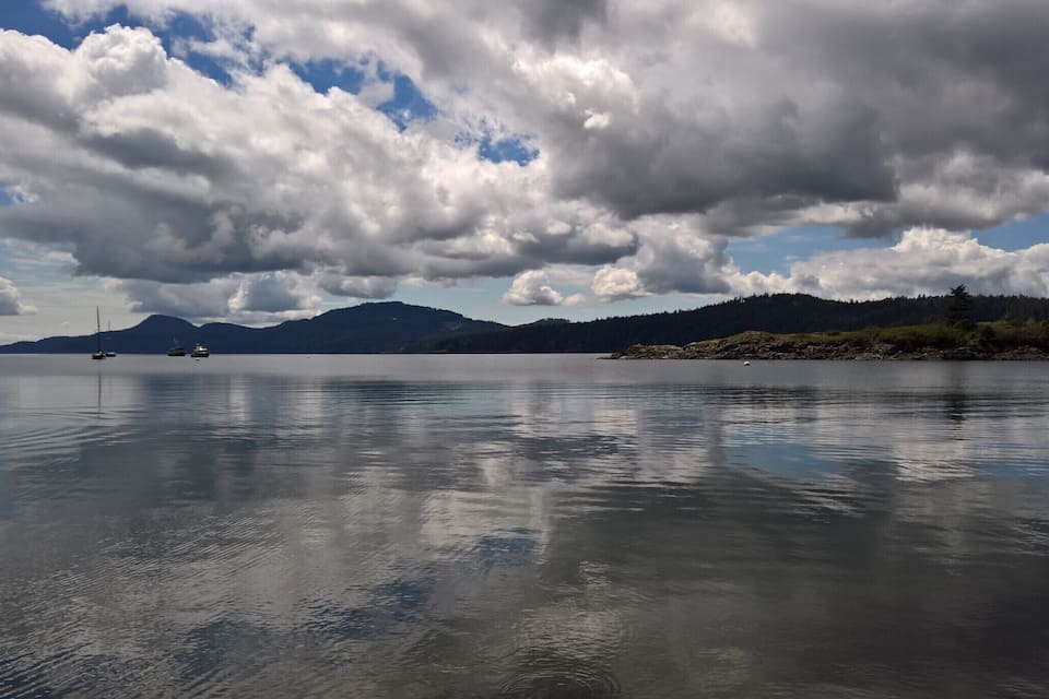 Un lago tranquilo con barcos, una isla boscosa y montañas a lo lejos bajo un cielo nublado.