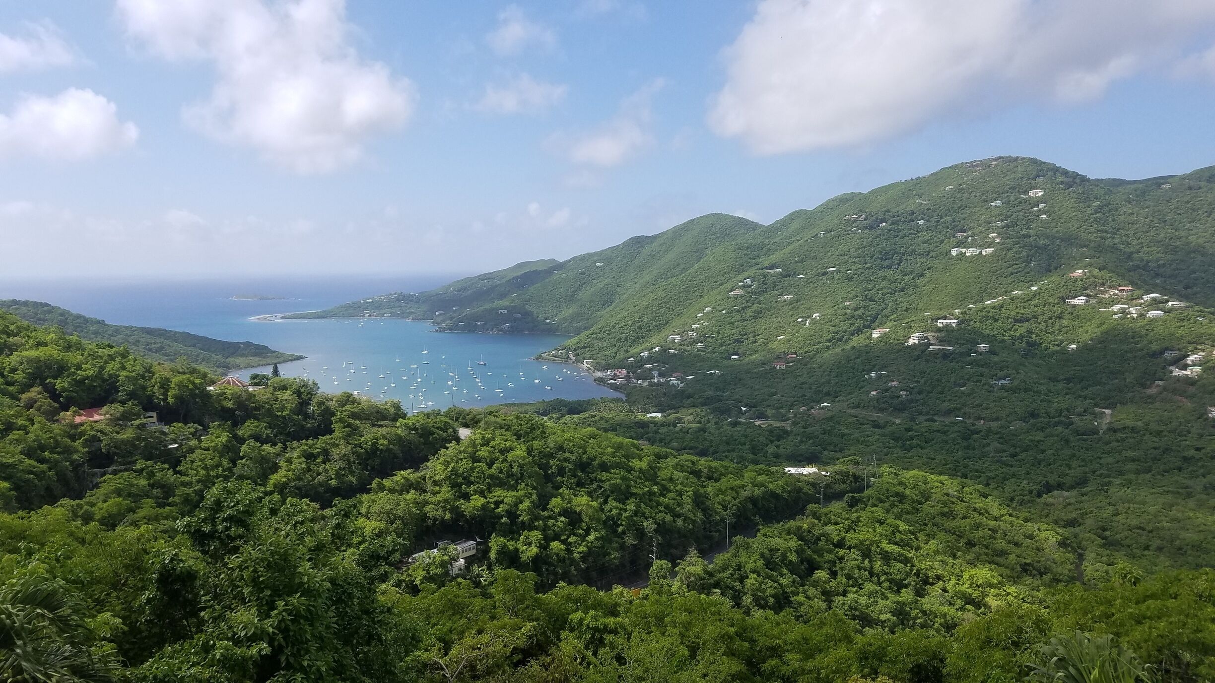 A coastal landscape with lush green hills, a bay with boats, and scattered buildings.