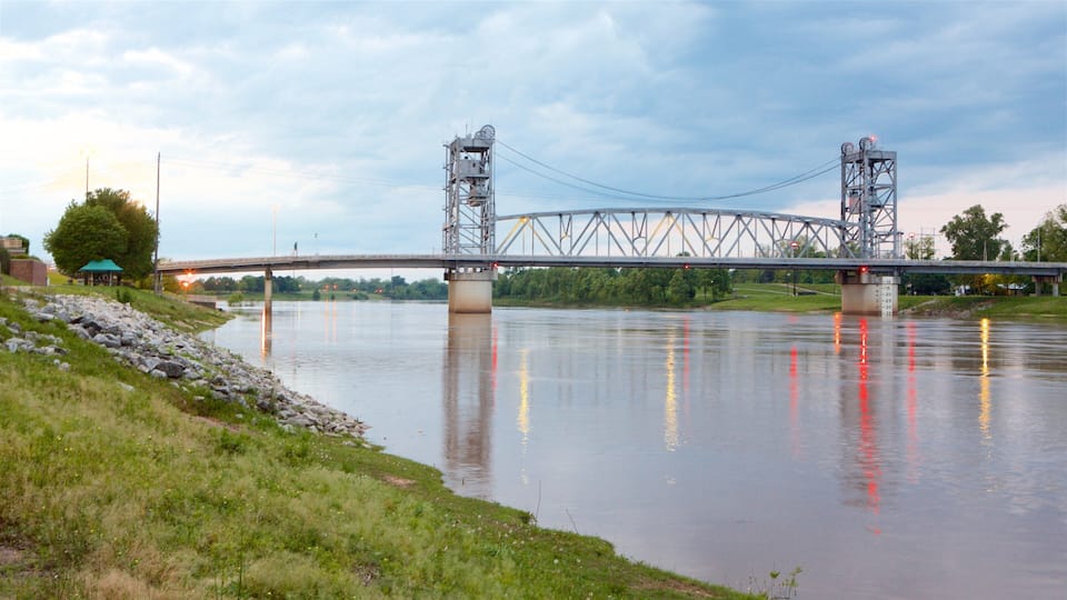 A steel truss bridge over a calm river with a grassy bank and a small building in the background.