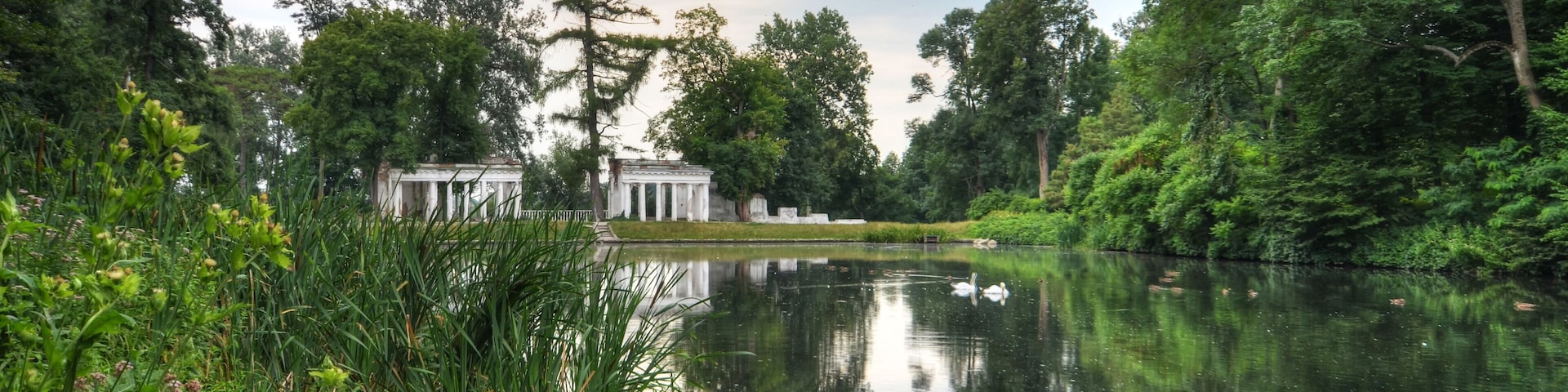 Um lago tranquilo com um edifício clássico ao fundo, rodeado por vegetação exuberante e árvores.