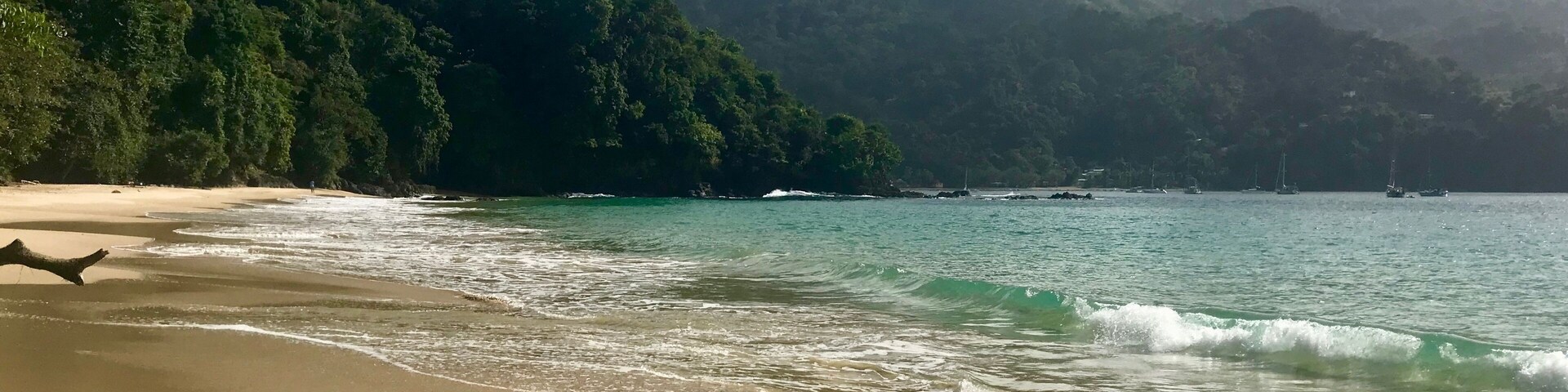 Uma praia com águas turquesa cristalinas, uma faixa de areia e colinas verdes exuberantes ao fundo.