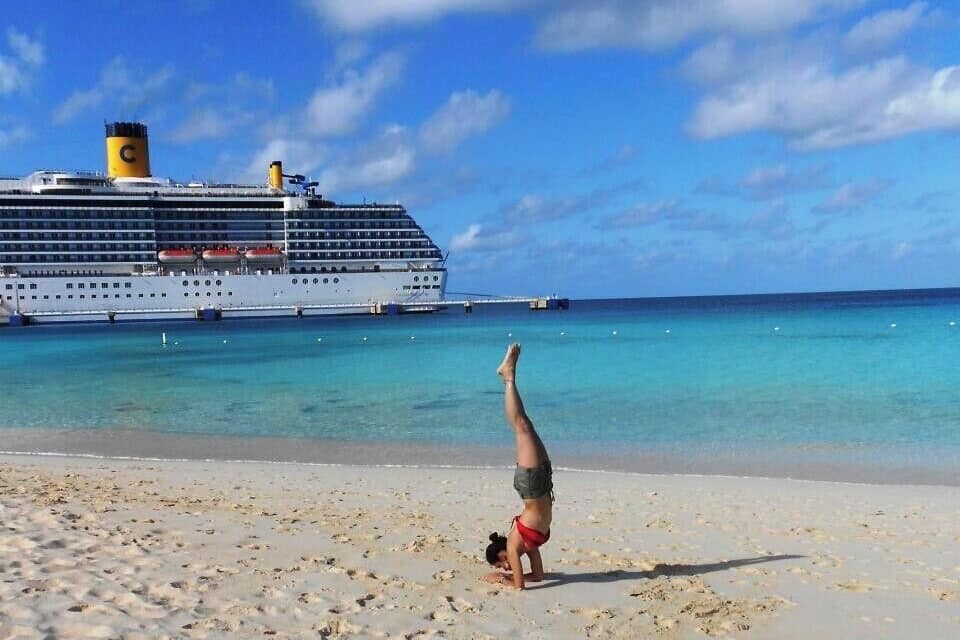 A cruise ship docked at a beach with clear turquoise water and a person performing a handstand on the sand.