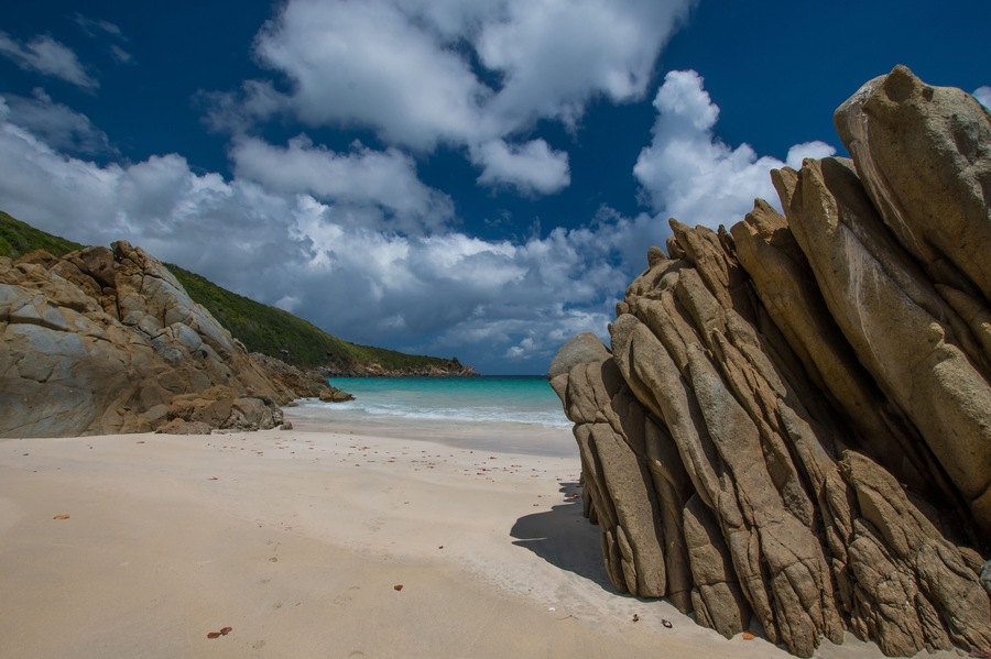 Uma praia com grandes formações rochosas, areal arenoso e céu azul claro com nuvens.