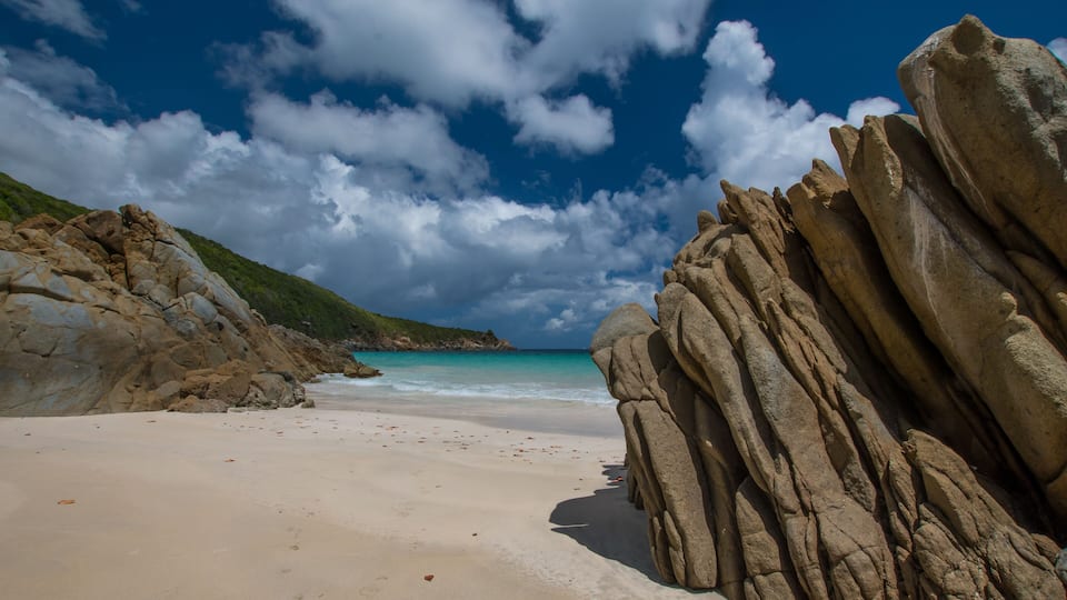 A beach with large rock formations, a sandy shore, and a clear blue sky with clouds.
