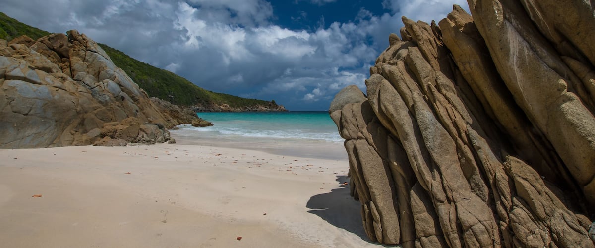 A beach with large rock formations, a sandy shore, and a clear blue sky with clouds.