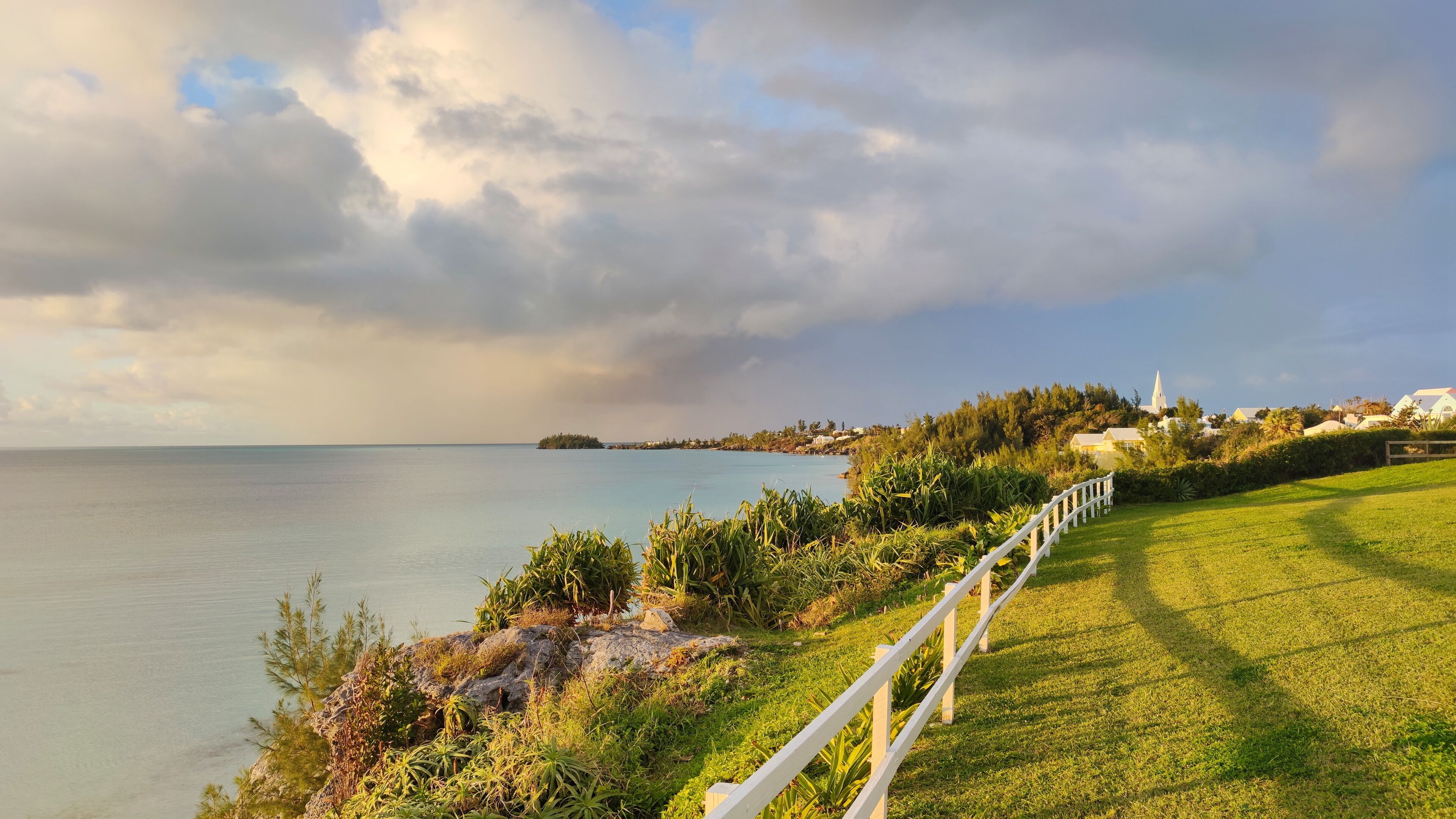A coastal landscape with a grassy area, a white fence, and a distant island with buildings and a church.