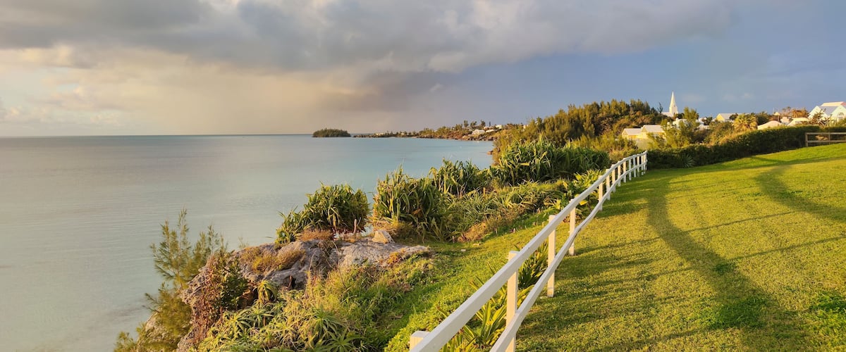 A coastal landscape with a grassy area, a white fence, and a distant island with buildings and a church.