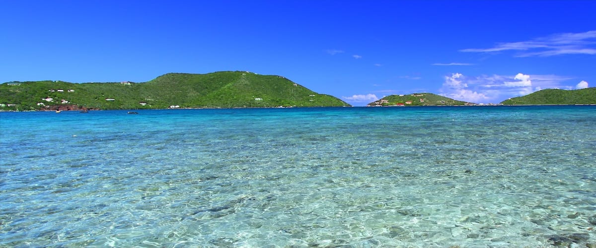 A clear turquoise sea with visible underwater rocks and two green hills in the distance.