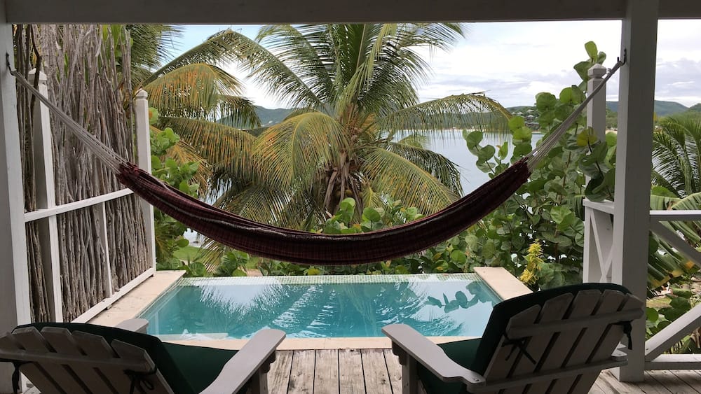 A wooden deck with a pool, two Adirondack chairs, and a hammock overlooking a tropical landscape with palm trees and a view of the water.