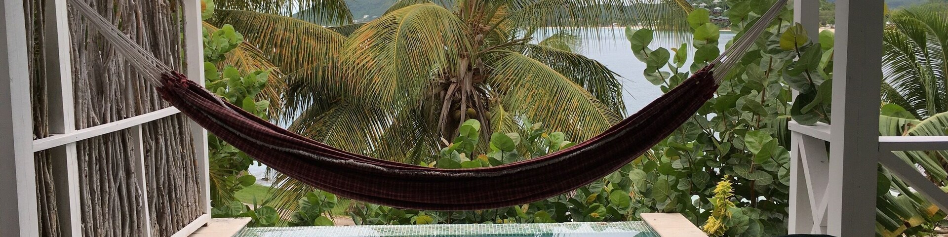 Um deck de madeira com piscina, duas cadeiras Adirondack e uma rede, com vista para uma paisagem tropical com palmeiras e uma vista da água.