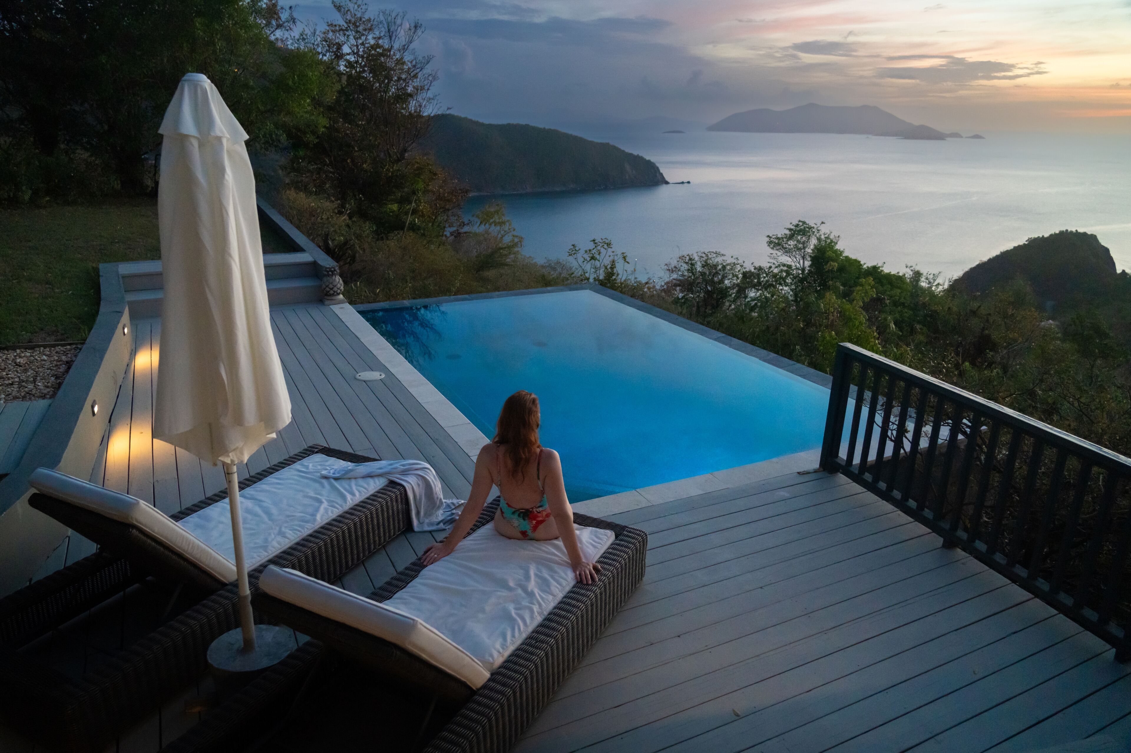 A woman relaxing on a deck chair by a pool with a view of the ocean and hills.