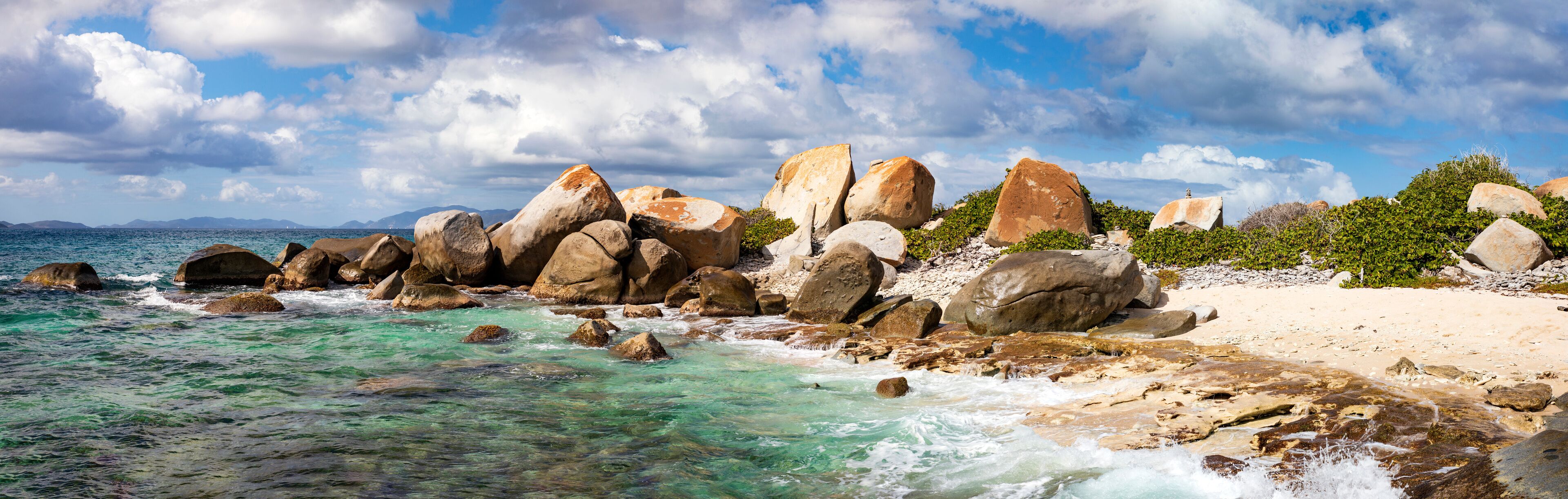A rocky shoreline with clear turquoise water, large boulders, and a sandy beach under a partly cloudy sky.