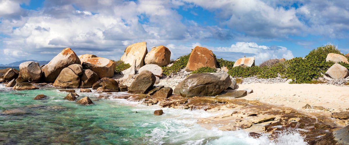 A rocky shoreline with clear turquoise water, large boulders, and a sandy beach under a partly cloudy sky.