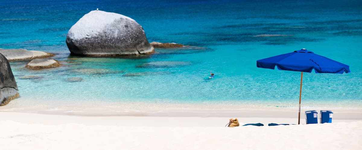 A beach with large rocks, clear turquoise water, and a blue umbrella.