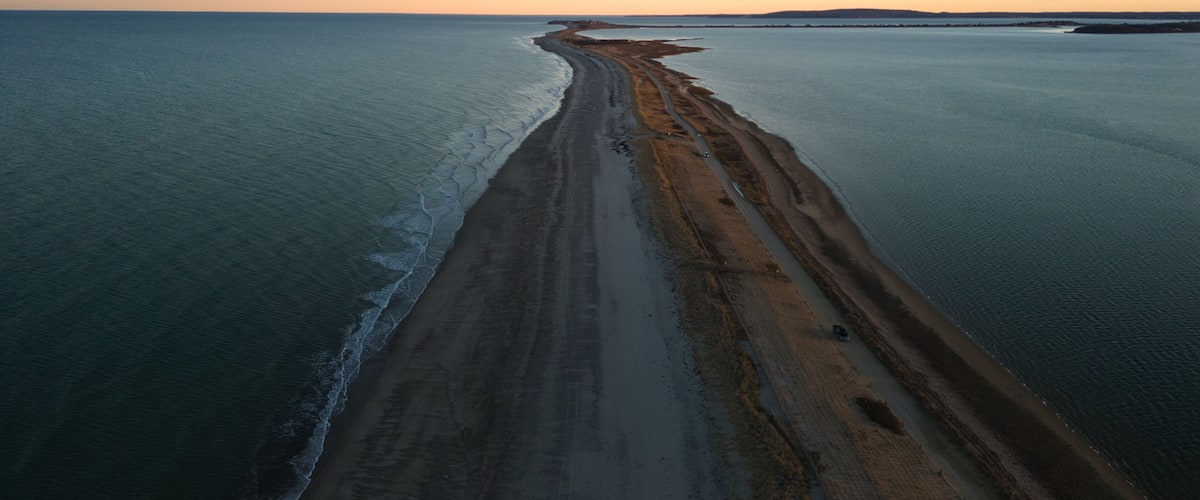 An aerial view of a long, narrow beach with a gentle curve, bordered by calm sea waters.