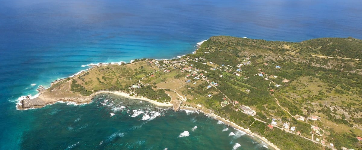 An aerial view of a coastal area with a beach, clear blue waters, and a hilly landscape with scattered buildings.