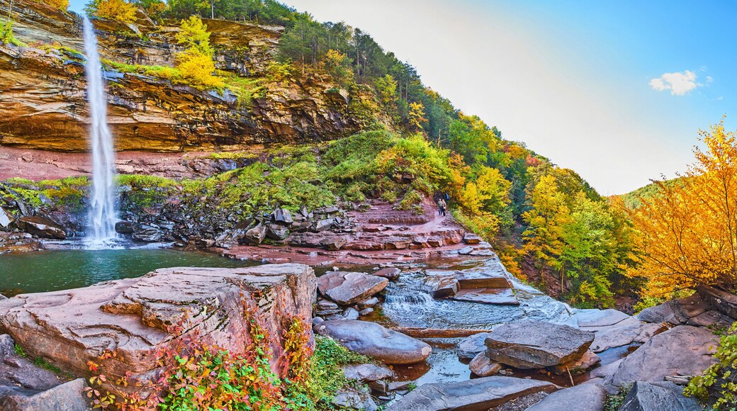 Wide panoramic view from below of waterfall over cliffs and fall foliage, hiking trail, and tourists