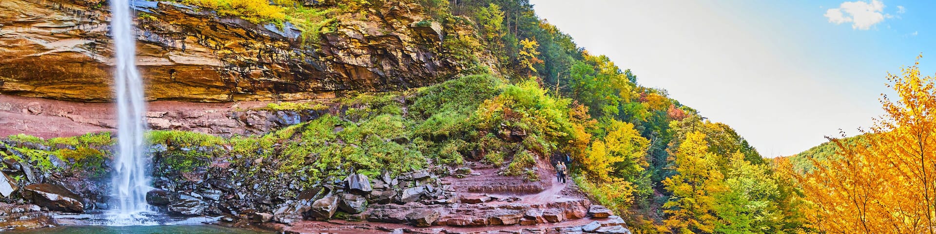 Wide panoramic view from below of waterfall over cliffs and fall foliage, hiking trail, and tourists