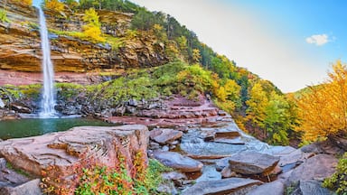 Wide panoramic view from below of waterfall over cliffs and fall foliage, hiking trail, and tourists
