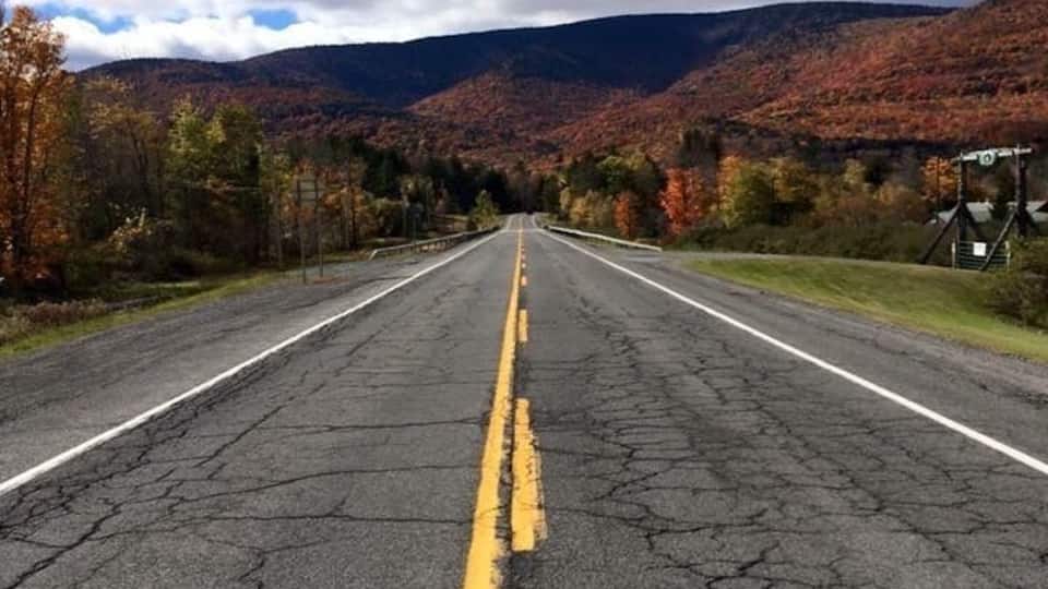 A road with a yellow center line, surrounded by autumn-colored trees and mountains in the distance.