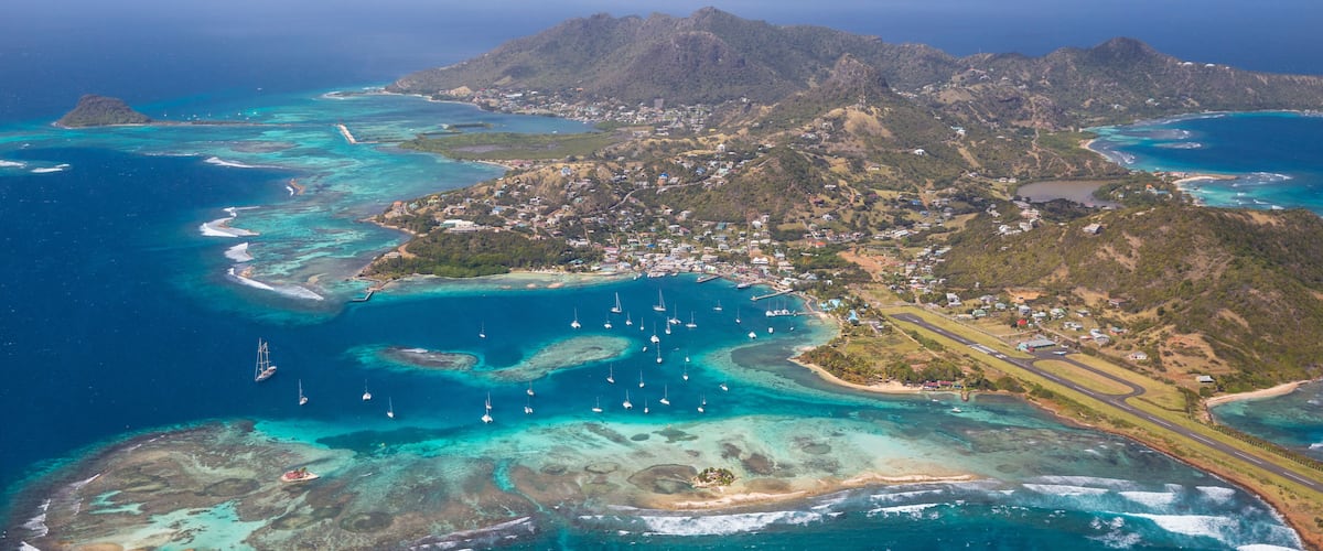 An aerial view of a coastal area with clear turquoise waters, a small island with a runway, and a mountainous backdrop.