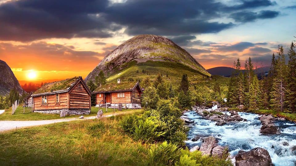 A wooden cabin by a fast-flowing river, surrounded by lush greenery and mountains under a sunset sky.