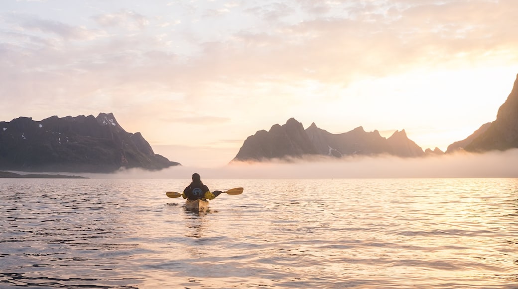 A kayaker in a calm lake surrounded by rugged mountains during sunrise or sunset.