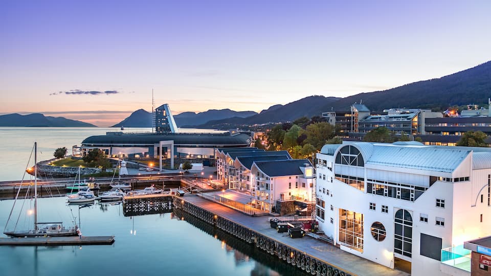 A waterfront marina with boats, a white building with a large circular window, and a backdrop of mountains and a clear sky.