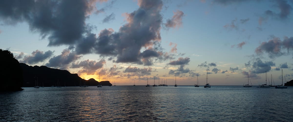 A sunset over a calm sea with sailboats and a mountainous coastline.