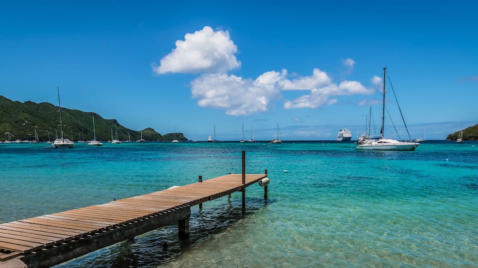 A wooden pier extending into turquoise waters with sailboats and a cruise ship in the background.