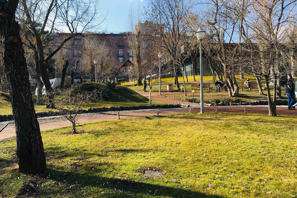A park with grass, trees, benches, and a building in the background.