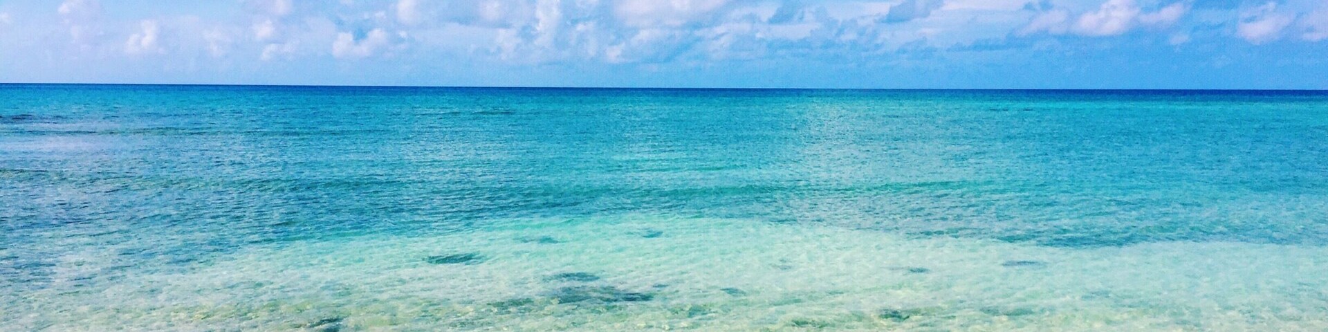 Mar turquesa translúcida, praia de areia branca e céu azul vibrante com nuvens fofas.