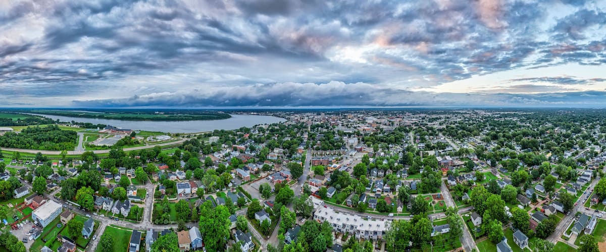 An aerial view of a town with a river, dense greenery, and cloudy skies.