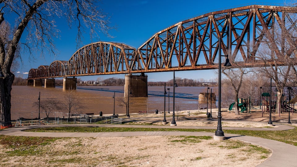 A large, rusted steel bridge over a river, surrounded by a park with trees and a playground.