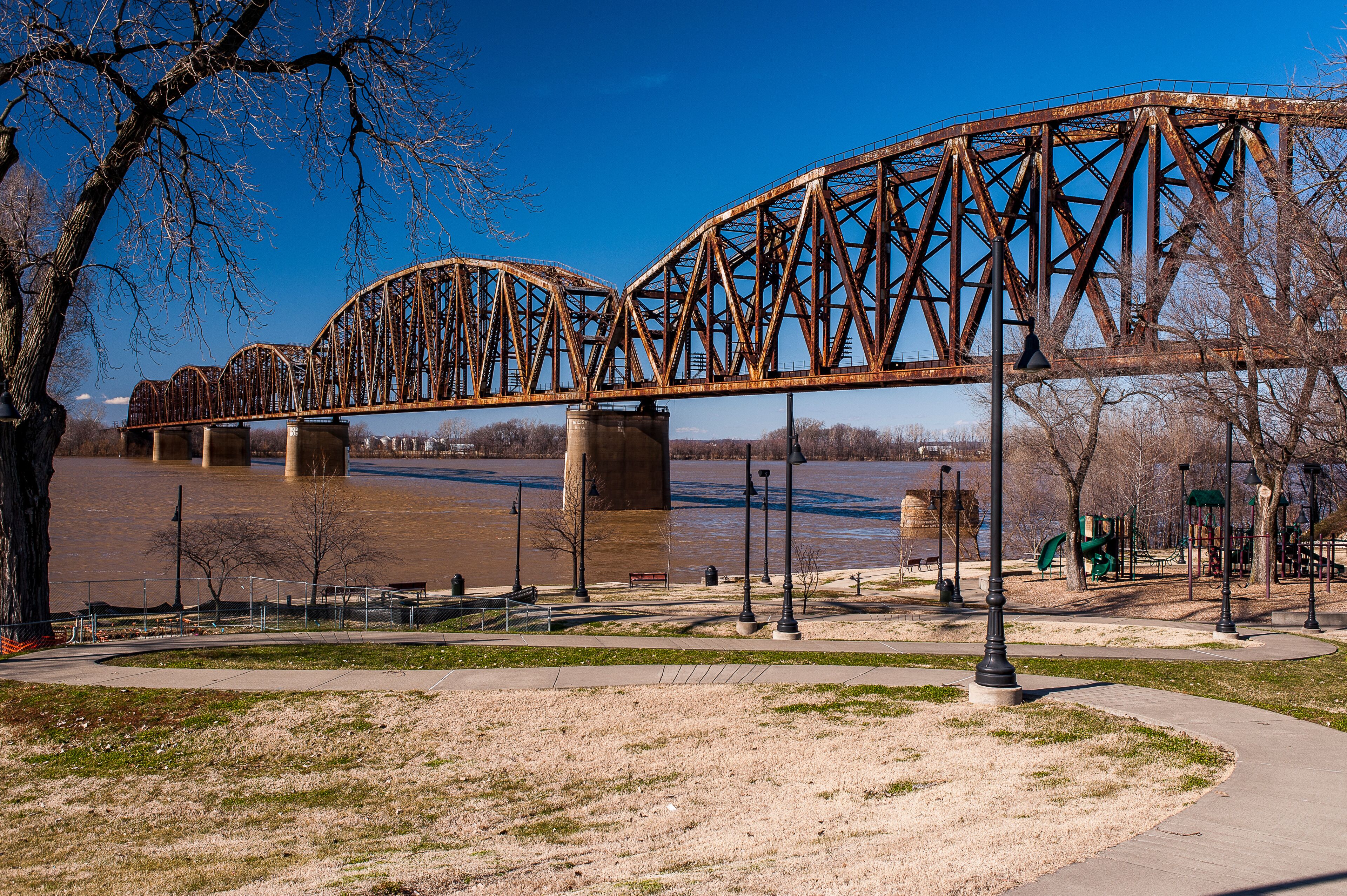 A large, rusted steel bridge over a river, surrounded by a park with trees and a playground.