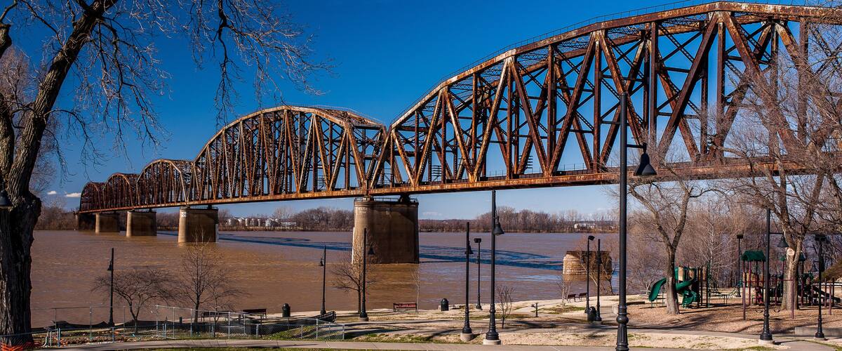A large, rusted steel bridge over a river, surrounded by a park with trees and a playground.