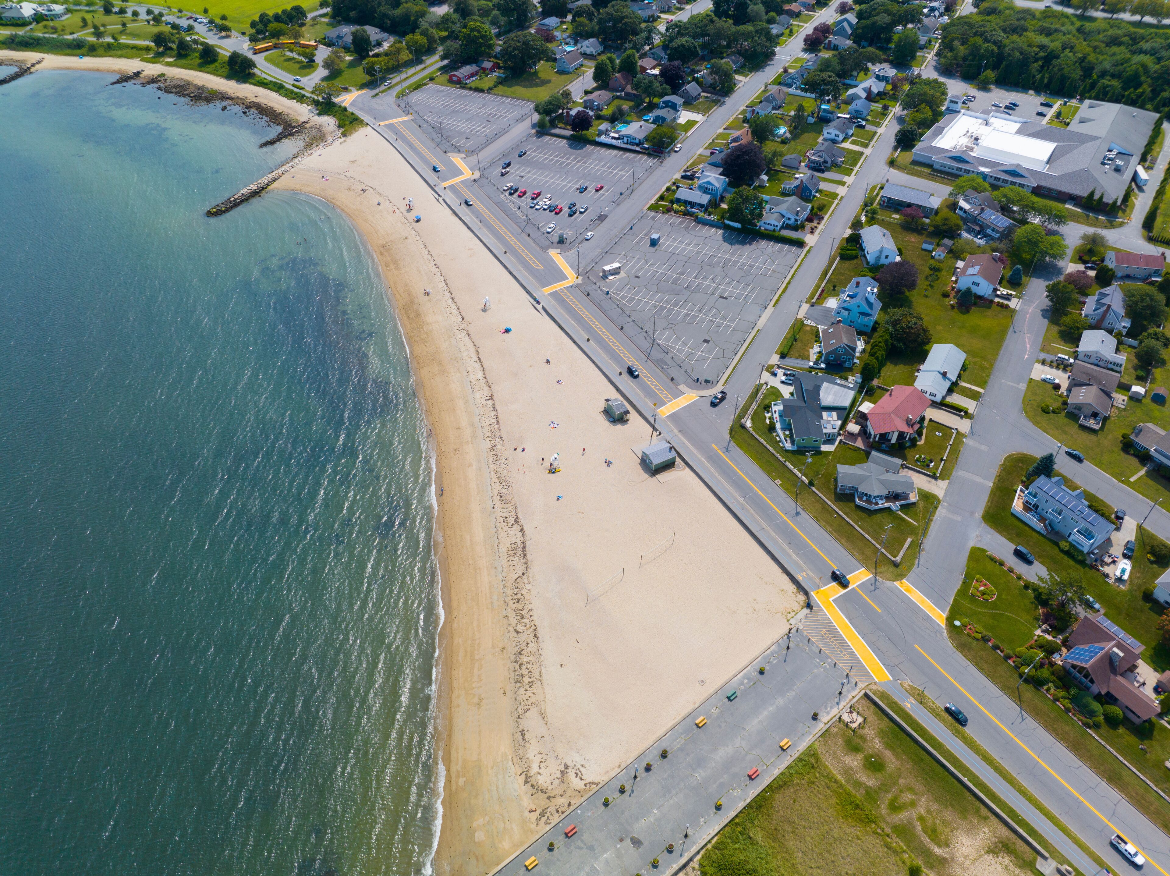 An aerial view of a coastal town with a beach, residential areas, and a parking lot.