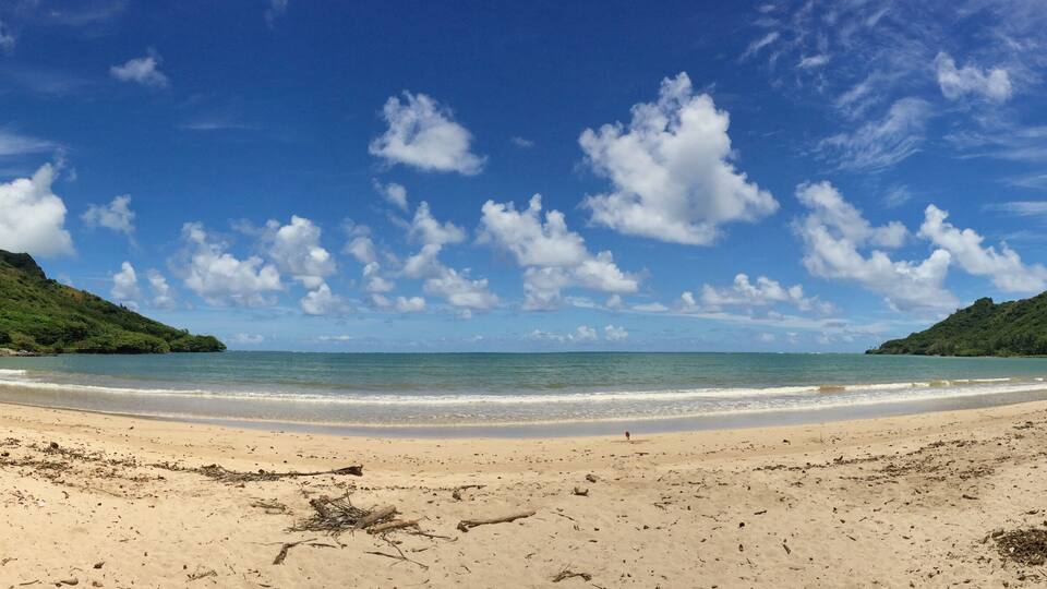A beach with clear blue water, sandy shore, and green hills in the background.