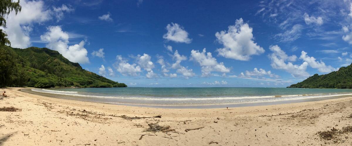 A beach with clear blue water, sandy shore, and green hills in the background.