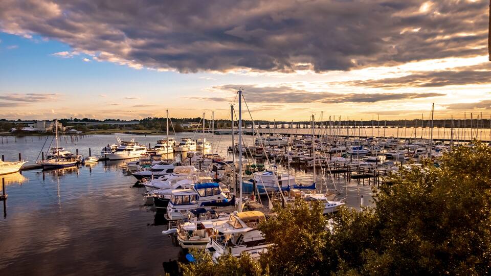 A marina with numerous boats docked, a bridge in the background, and a cloudy sky.