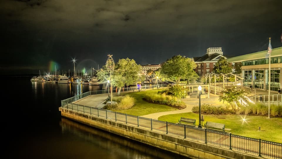 A waterfront promenade at night with a calm river, illuminated trees, benches, and a distant building.
